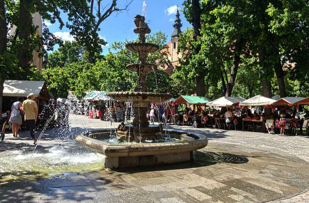 „Englisch lernen in Ungarn – internationale Begegnungen auf einem lebendigen Marktplatz mit Springbrunnen und bunten Marktständen als Symbol für Sprachaustausch und Integration.