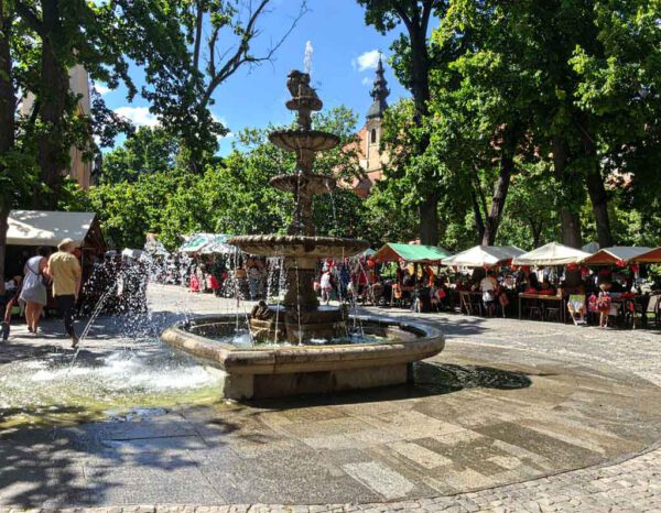 „Englisch lernen in Ungarn – internationale Begegnungen auf einem lebendigen Marktplatz mit Springbrunnen und bunten Marktständen als Symbol für Sprachaustausch und Integration.