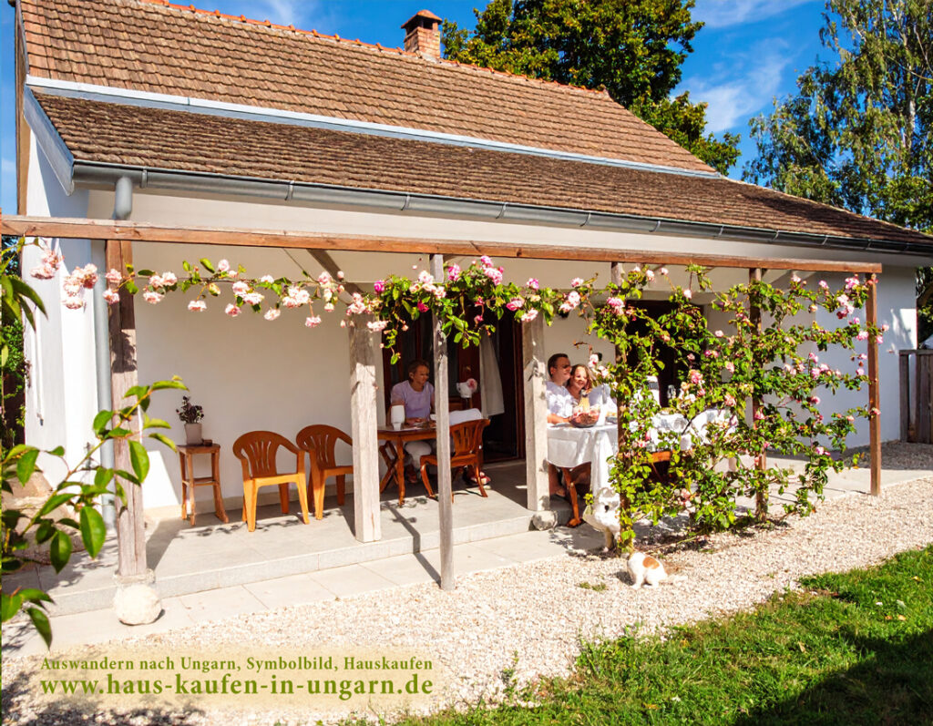 Menschen sitzen bei einem gemeinsamen Essen auf einer blumengeschmückten Terrasse unter einer Pergola vor einem weiß gestrichenen Landhaus – Symbolbild für das Auswandern und Haus kaufen in Ungarn