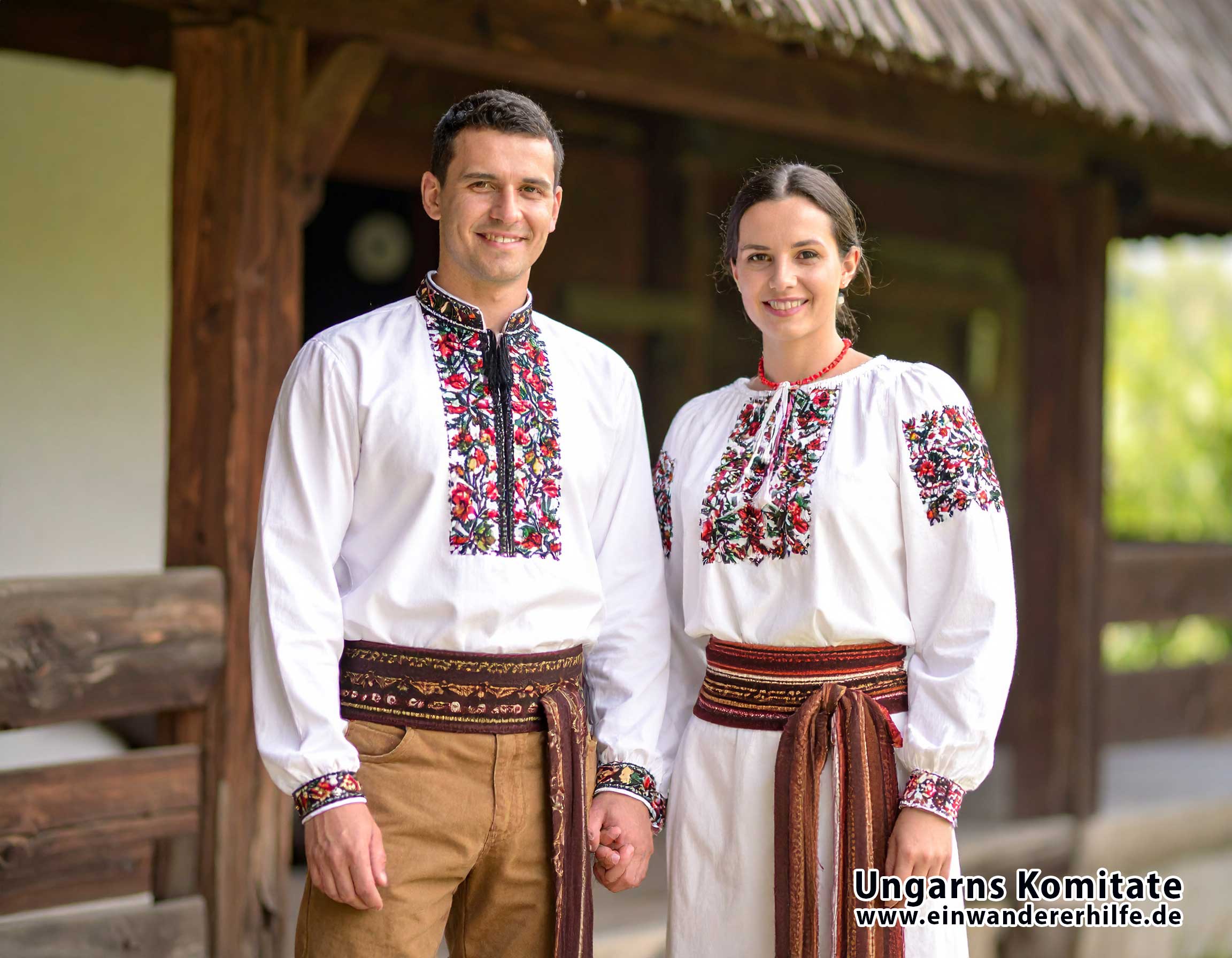 Ein ungarisches Ehepaar in farbenfroher Folklorekleidung steht stolz vor einer traditionellen Lodge. Die Frau trägt eine bestickte Bluse, der Mann ein Hemd mit weiter Hose und breitem Gürtel. Gemeinsam verkörpern sie die gelebte Tradition, Gastfreundschaft und das ländliche Erbe Ungarns.
