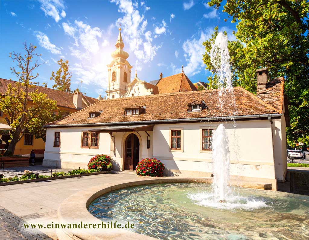 Bild symbolisiert einen Ortskern. Vép in Ungarn mit Kirche, Brunnen und gepflegtem Garten bei sonnigem Wetter