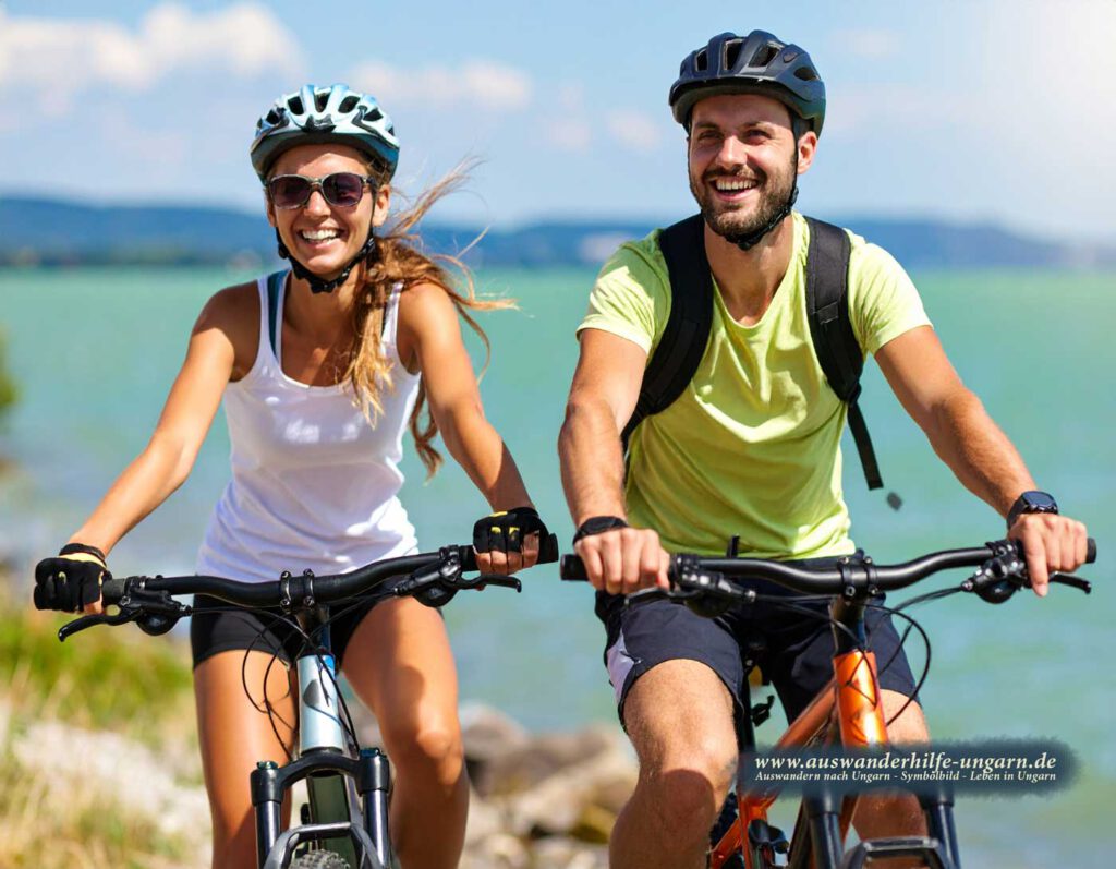 Zwei hellhäutige europäische Radfahrer auf dem Balaton-Radweg, lachend, glitzerndes Wasser, sonnig.