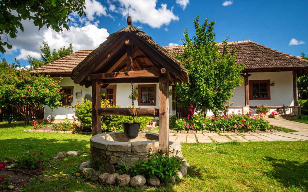 Traditionelles ungarisches Haus mit Strohdach und Steinbrunnen in Gencsapáti, umgeben von grüner Natur und Sommerhimmel - Symbolbild, Marion Schanné