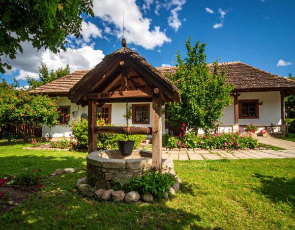 Traditionelles ungarisches Haus mit Strohdach und Steinbrunnen in Gencsapáti, umgeben von grüner Natur und Sommerhimmel - Symbolbild, Marion Schanné