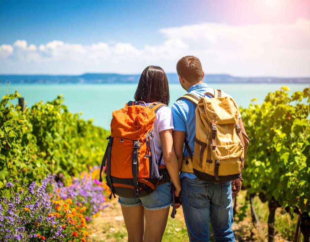 Lächelndes Paar mit Rucksäcken umarmt sich auf einem Wanderweg durch blühende Weinberge mit Blick auf den Kis-Balaton in Ungarn