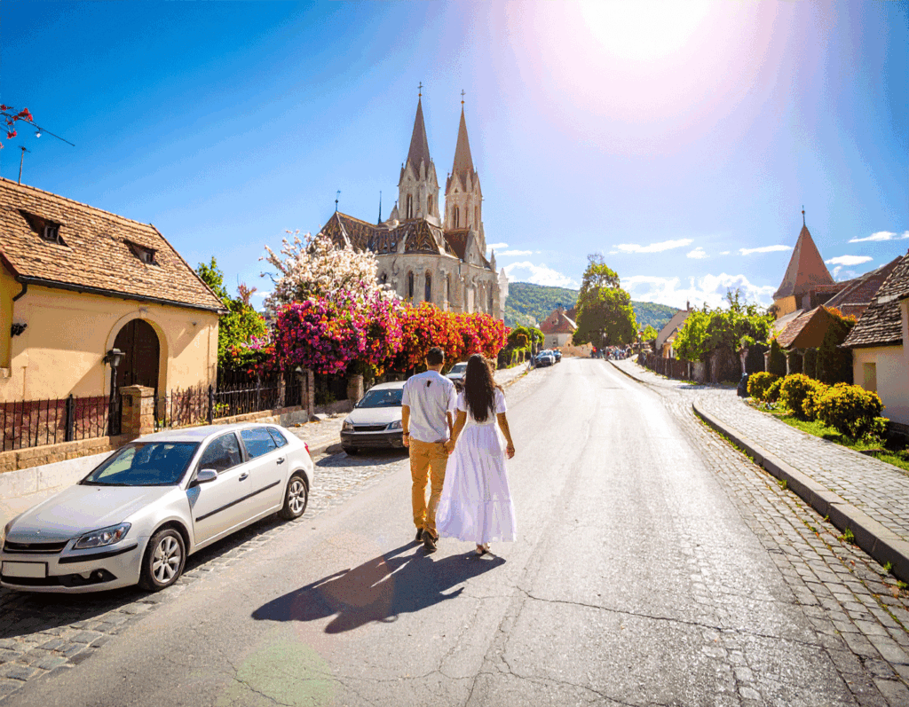 Ein Paar in weißer Sommerkleidung spaziert auf einer sonnigen Dorfstraße; links traditionelle Häuser, rechts Gärten, vorne eine Kirche mit zwei Türmen – Symbolbild, nicht Táplánszentkereszt