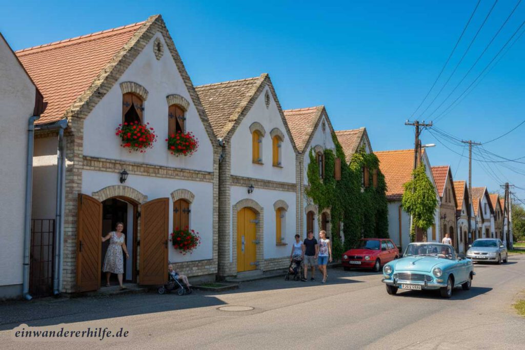 Bacs Kiskun - Auswandern nach Ungarn: Ein sonniger Sommertag in einem ungarischen Dorf mit traditionellen Häusern, einer Frau, die aus einem Haus mit blühenden Geranien tritt, und Menschen, die auf der Straße spazieren gehen und in Autos fahren.