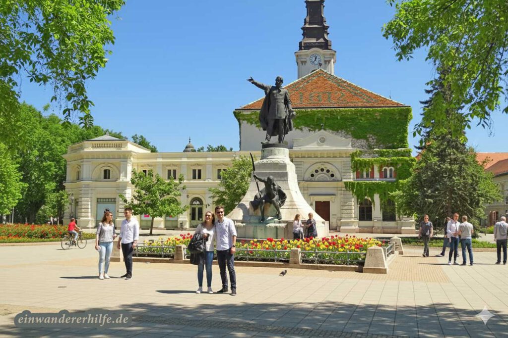 Glückliche Menschen genießen den Frühling in einer belebten Stadtszene vor einem Denkmal und einem historischen Gebäude mit Kirchturm im Komitat Bács-Kiskun.