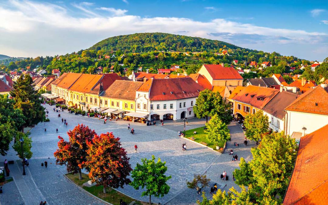 Kecskemet vibes - SymbolBild einer ungarischen Altstadt mit Marktplatz, roten Dächern und Blick auf grüne Hügel im Hintergrund.