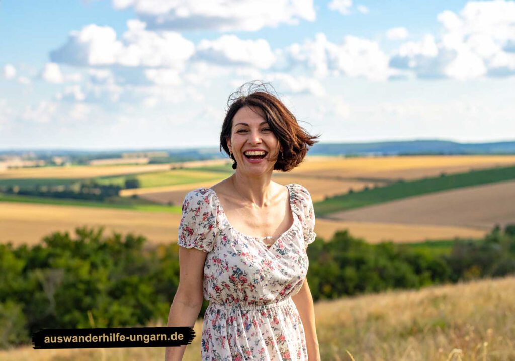 Siklos Umgebung: Fröhliche Frau in Blumen-Sommerkleid lacht herzlich auf einer Wiese mit weitem Blick über die ungarische Landschaft – Symbol für Lebensfreude, Mut und Auswanderung.