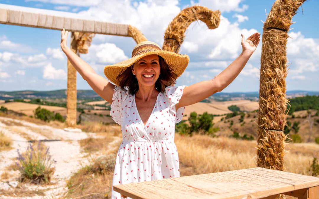 Siklos im Sommer. Lachende Frau in weißem Sommerkleid und Strohhut hebt die Arme in die Luft, sonnige Landschaft in Ungarn im Hintergrund – Symbol für Freiheit, Mut und Auswandern.