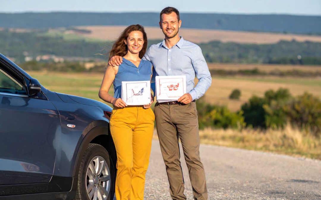 Ein lächelndes Paar steht neben einem Auto auf einer Landstraße in Ungarn und hält stolz ihre Dokumentenmappen in der Hand. Das Foto symbolisiert den erfolgreichen Erwerb des Führerscheins in Ungarn 2025 und den selbstbewussten Start in die mobile Freiheit.