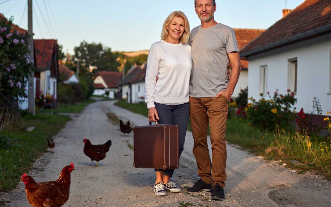 Ein sympathisches Paar um die 50 steht mit einem Koffer auf einer sonnigen Dorfstraße in Ungarn, während Hühner über den Weg laufen. Das Foto symbolisiert das Auswandern nach Ungarn – den Schritt in ein ruhiges, einfaches und naturverbundenes Leben auf dem Land.