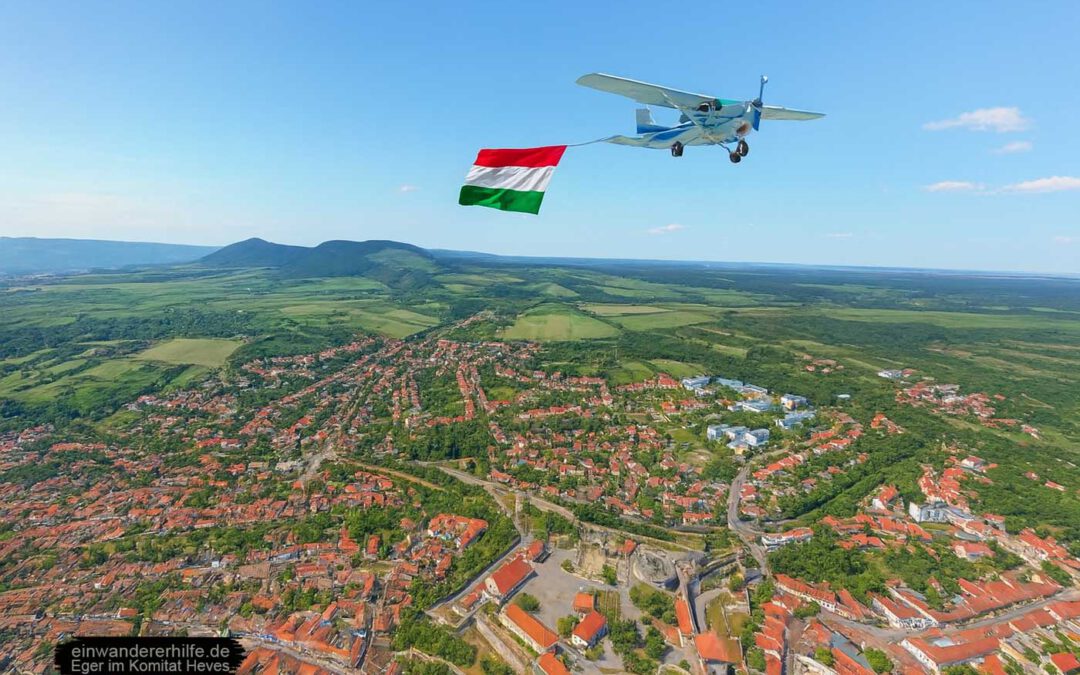 Ein Kleinflugzeug mit ungarischer Flagge fliegt über die Stadt Eger im Komitat Heves – Blick auf Dächer, Landschaft und Weinregion.