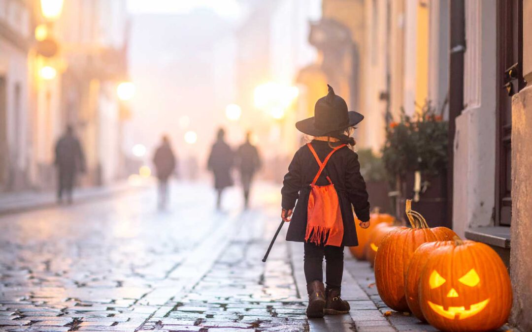 Halloween in Ungarn: Ein kleines Mädchen im Hexenkostüm mit orangefarbenem Umhang geht in der Abenddämmerung durch eine gepflasterte Altstadtstraße; am Straßenrand stehen leuchtende Kürbisse und verbreiten herbstliche Stimmung.