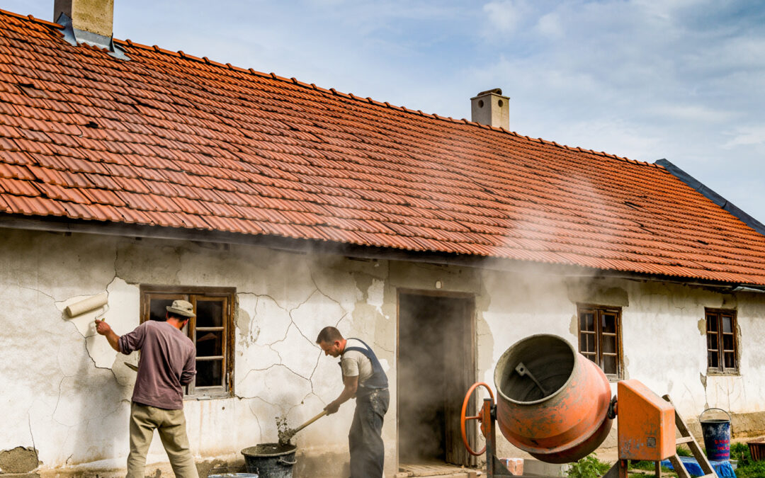 Handwerker in Ungarn renovieren ein altes eingeschossiges Bauernhaus mit rotem Ziegeldach. Einer streicht die Fassade mit der Rolle, der andere mischt Zement neben einem Betonmischer. Staub und Sonnenlicht schaffen eine authentische Dorfszene, wie sie für Renovierungen auf dem ungarischen Land typisch ist.