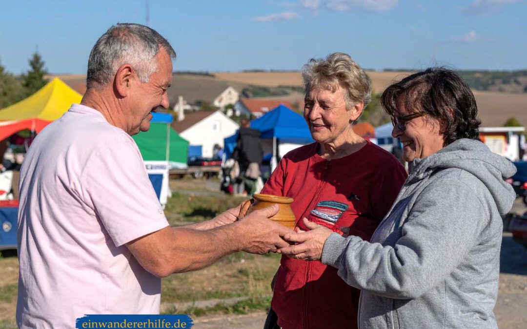 Ein älterer Mann überreicht zwei Frauen auf dem Wochenmarkt wie damals in Mezőtúr in der ungarischen Tiefebene ein handgefertigtes Tongefäß; freundliche Gesichter, bunte Marktstände und weite Felder im Hintergrund.
