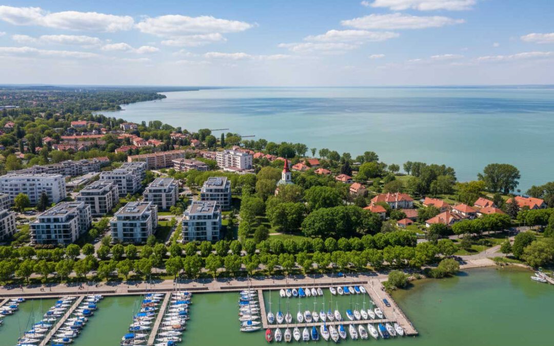 Blick auf den Yachthafen, die moderne Uferpromenade und die roten Dächer der Stadt am Balaton, eingebettet in grüne Bäume und blaues Wasser unter klarem Sommerhimmel.
