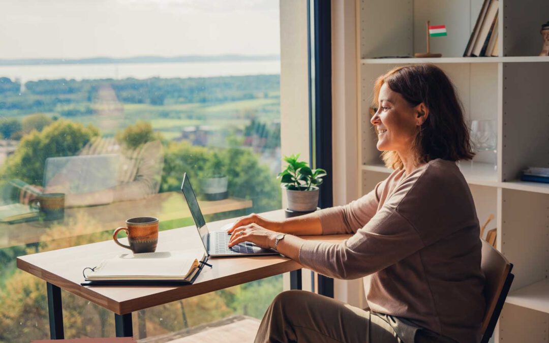 Deutsche Frau um die 50 arbeitet lächelnd im HomeOffice in Ungarn an einem Laptop, vor großer Fensterfront mit Blick über eine grüne Landschaft und einen See.