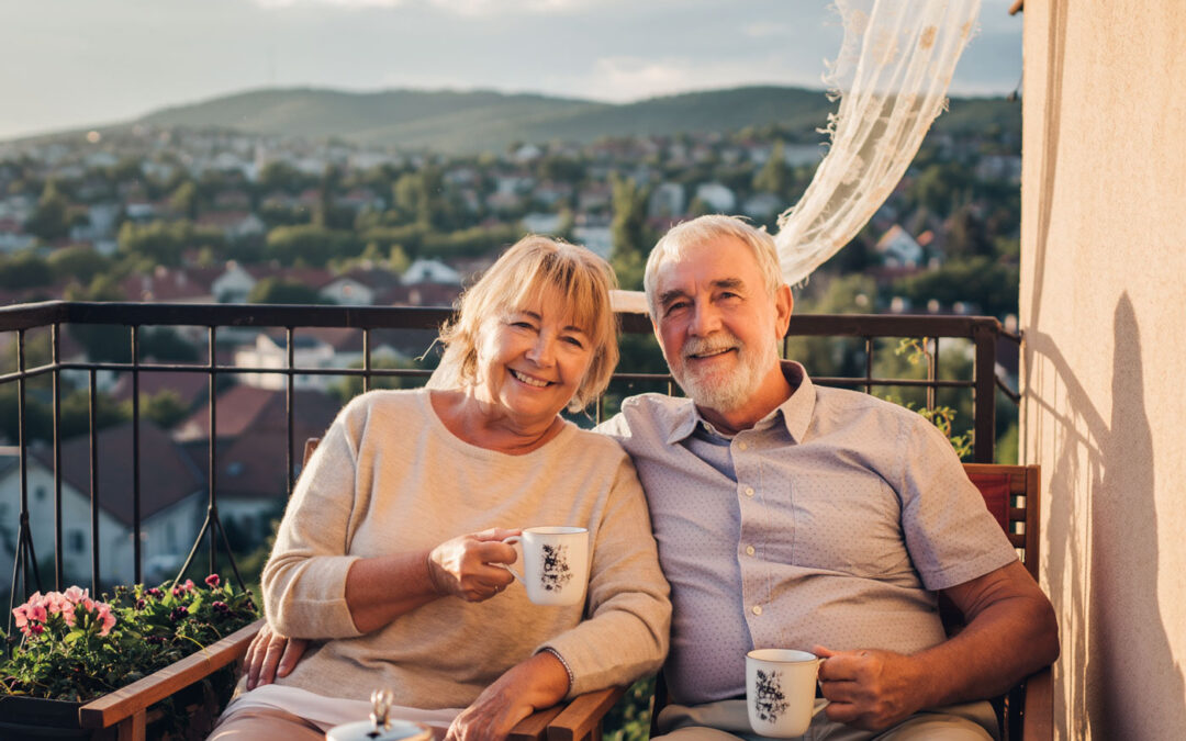 Lächelndes Seniorenpaar sitzt mit Kaffeetassen auf einem Balkon mit Blick über eine ungarische Stadt und genießt sein neues Leben nach der Auswanderung nach Ungarn.